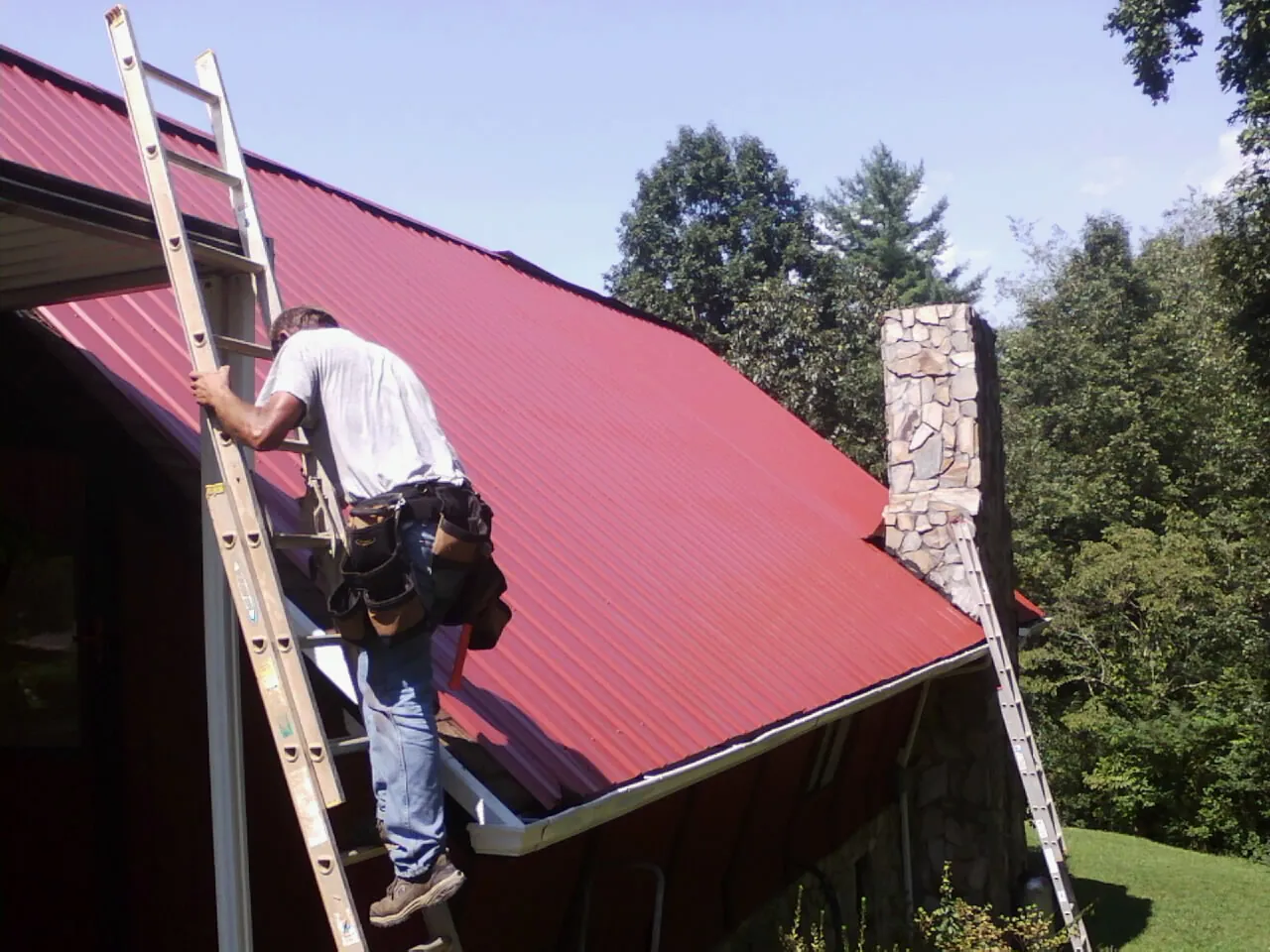 Roofer on a ladder beside a red metal roof and stone chimney — Soffit & Fascia Repair by Roofing Brothers in Rescue